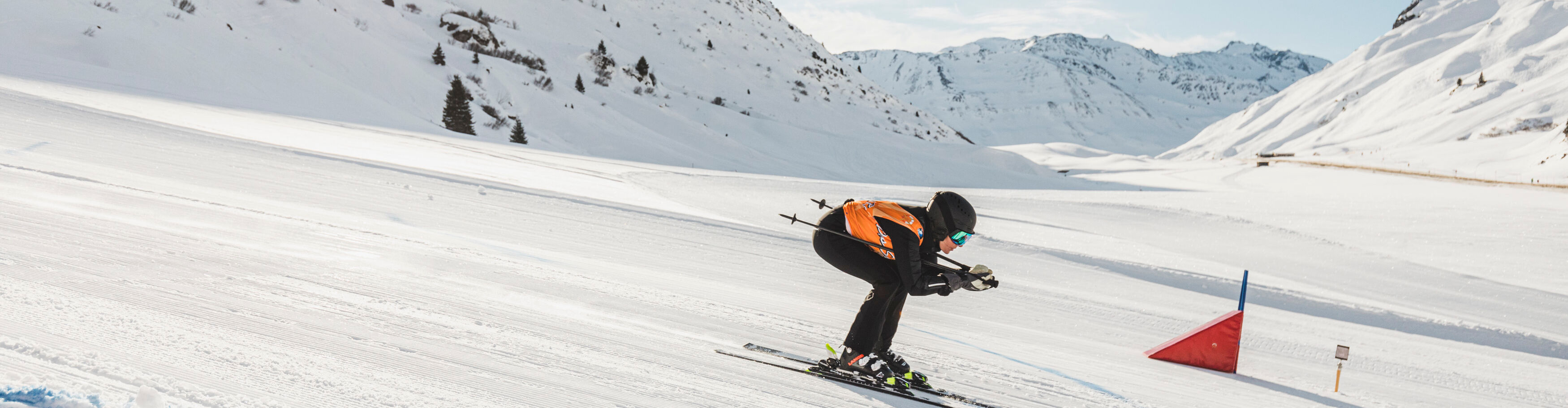 Im Vordergrund fährt ein Skifahrer in Hocke ein Rennen runter und im Hintergrund ist eine verschneite Bergkulisse zu sehen. 